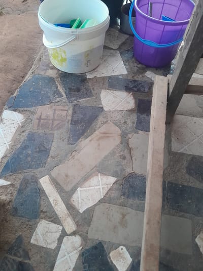 The scene is located in a utility area in Ifo, Nigeria, observed from an elevated perspective. The ground is paved with an irregular mosaic of broken ceramic tiles and stone fragments, primarily in shades of dark grey, light grey, and white, set within a light brown cementitious matrix; some white tile fragments display etched or painted cross-hatch patterns. In the upper left, a white plastic bucket, exhibiting yellow-green stains and some legible text including "PT Maligana," contains a blue plastic scoop, a green sponge, and a clear-green plastic item. To its right, a purple plastic bucket with a light blue handle rests on the tiled surface, holding a blue plastic tool and a thin, dark rod; a small amount of liquid is visible on the ground around its base. Partially obscured metallic pots are present in the background between the two buckets. A light-colored wooden beam extends vertically along the right side of the frame. No individuals are present.