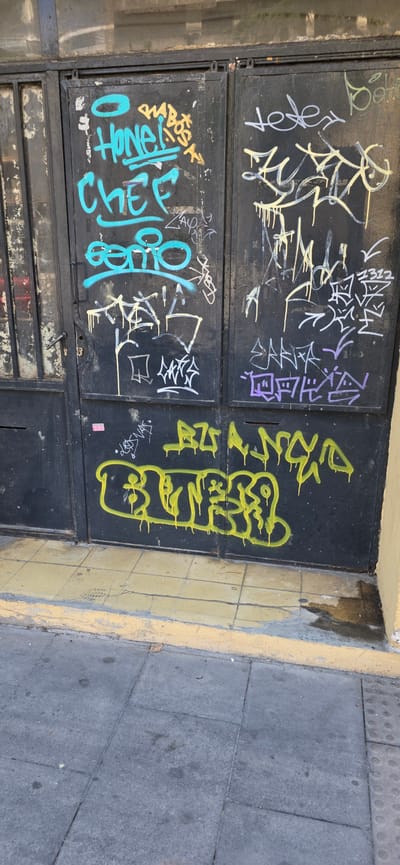 The image shows a close-up, eye-level view of a black metal gate covered in graffiti. The gate appears to be old and weathered, with some rust and chipped paint visible. The graffiti is a mix of tags and bubble letters in various colors, including blue, yellow, purple, and white. Some of the graffiti is dripping, suggesting it was recently applied.
In the lower portion of the image, a yellow tiled sidewalk is visible, with a grey concrete sidewalk in front of it. The overall scene suggests an urban environment, possibly an alleyway or the entrance to a building. There are no people visible in the image.
The location of Huesca, Spain, is not explicitly indicated in the image.