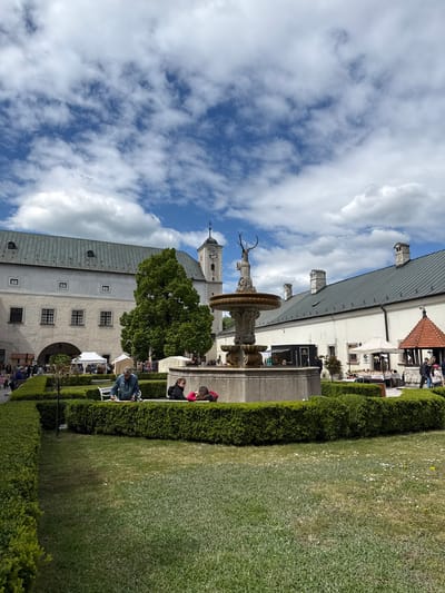 The image shows a view of Červený Kameň Castle in Častá, Slovakia, featuring a fountain with a statue of a deer, lush green lawns, and manicured hedges. People are seen sitting on benches and walking around the courtyard. The architecture of the castle and surrounding buildings is visible, including a clock tower. The sky is blue with scattered white clouds.