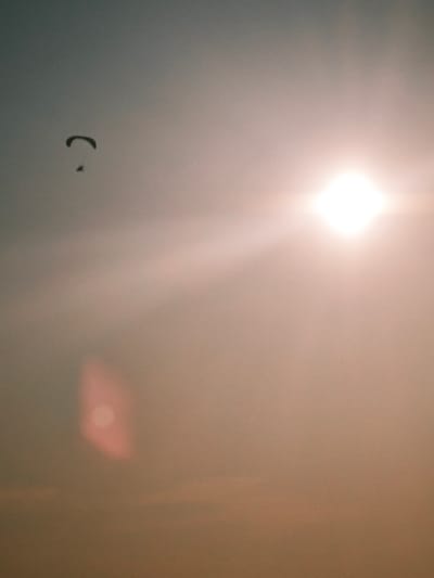 The image depicts a paraglider in silhouette against a hazy sky, with the sun prominently visible as a bright, square-shaped lens flare. The paraglider, a small dark shape, is positioned in the upper left quadrant of the image, with its canopy curved and a smaller figure visible beneath it. The sky is a gradient of soft colors, transitioning from a muted grey-blue at the top to a warm, peachy tone towards the bottom, likely indicating sunset or sunrise. A large, bright sun, emitting rays of light, is located in the upper right portion of the frame. A reddish, out-of-focus light flare is visible in the lower left quadrant. The overall atmosphere is hazy and somewhat dreamlike due to the atmospheric conditions and the lens flare. There is no indication of the city of Lesosibirsk, Russia, in the image.