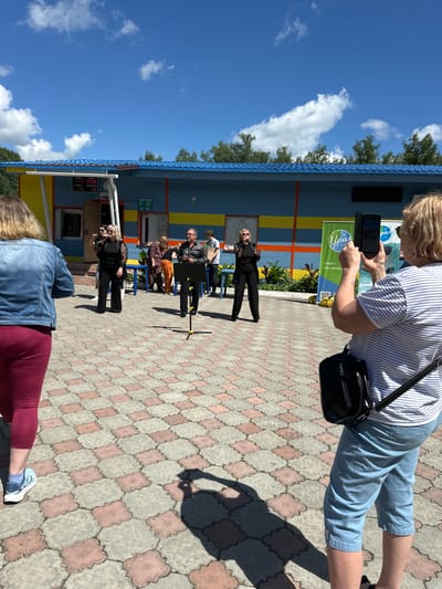 The image depicts an outdoor performance or event in front of a brightly colored building, possibly a public facility or a resort in Duvankul', Russia. The building has a blue roof and yellow and blue striped walls. In the foreground, a woman with blonde hair, wearing a striped t-shirt and blue shorts, is holding up a smartphone to record the scene. She is positioned to the right of the frame. In the mid-ground, two women in black outfits and sunglasses are singing into microphones, standing near a music stand. Behind them, a man in a dark shirt and pants is also singing into a microphone, with a music stand in front of him. Several other individuals are present in the background, some appearing to be part of a group or audience. The ground is paved with interlocking hexagonal and quatrefoil-shaped tiles in shades of pink and gray. The sky is blue with scattered white clouds, and trees are visible beyond the building. A green banner with the logo "Урал-это санаторий" (Ural - this is a sanatorium) is visible behind the performers. The overall scene suggests a sunny day and a public gathering or entertainment event.