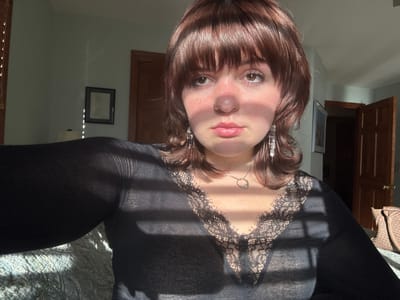 An upside-down view of a fair-skinned woman with bangs and shoulder-length brown hair. She wears a black top with lace trim at the neckline and a delicate necklace. Sunlight casts shadows across her face and clothing. A partially open wooden door and framed picture are visible in the background, along with a glimpse of a patterned bed covering. The room appears to be a bedroom or living space.
