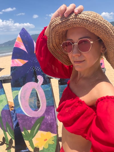 A woman with blonde hair is standing on a beach in Quy Nhon Ward, Vietnam. She is wearing a red off-the-shoulder top, sunglasses, and a straw hat. She is holding her hat with her left hand. In the background, there are two surfboards with colorful painted designs, the ocean, and mountains. The sky is blue with some clouds. The sand is light brown.