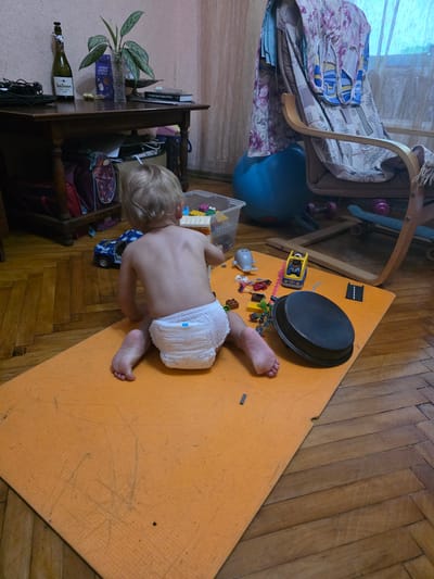 A toddler wearing a diaper sits on an orange mat on a wooden floor. Toys, including LEGO bricks, a toy car, and a small grey airplane, are scattered around the child. In the background, a wooden table holds a bottle, a plant, and books. A chair draped with fabric and a large blue exercise ball are also visible. The scene appears to be in an indoor residential setting.
