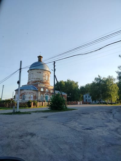 The image captures a view of a brick church with a large dome and a smaller dome, both topped with crosses, in the village of Pyrkino, Russia. The church appears aged, with visible wear on its brickwork and peeling paint on the main dome. A fence encloses the church grounds, and a few trees are visible in the foreground and background. To the right of the church, a two-story building with a light-colored facade and blue window frames stands amidst a cluster of deciduous trees. Overhead, multiple power lines stretch across the clear blue sky, with a few birds visible in flight. The ground in the foreground is a dirt and gravel road with some visible potholes. The overall scene suggests a rural setting on a sunny day.