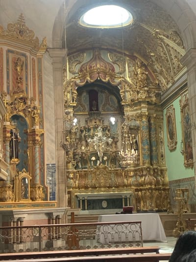 The image displays the highly ornate Baroque interior of the Igreja e Convento de São Francisco (Church and Convent of Saint Francis), located in the Pelourinho neighborhood of Salvador, Bahia, Brazil.
The scene is dominated by the elaborate main altarpiece, which is extensively covered in gilded woodcarvings (talha dourada) and features intricate scrolling, volutes, and statuettes. Teal-blue accents are integrated into the gilded framework. A large crucifix with a figure of Christ is centrally positioned above a white-draped altar table. A dark, possibly liturgical, book rests on the altar table. Numerous hanging chandeliers with crystal-like elements are suspended around the main altar, contributing to the illumination.
To the left of the main altar, a side altar structure, equally resplendent with gilded carvings and blue accents, houses statues of two robed figures, likely Franciscan friars. The ceiling is vaulted and richly decorated with gold ornamentation, featuring a prominent circular skylight (oculus) that provides natural light. The right wall, painted a light green, displays framed religious paintings.
In the foreground, sections of dark wooden pews are visible, along with a decorative metal railing separating the nave from the chancel. A person's dark hair is partially visible in the bottom right corner, indicating a viewer seated in a pew. The environment is one of extreme opulence, meticulously crafted and reflective of historic religious artistry. No explicit actions or interactions are depicted beyond the static arrangement of the church interior.