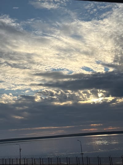 The image depicts a wide, static view of a dusky sky filled with clouds, over a calm body of water. The sun is setting or has recently set, casting a warm, golden glow through breaks in the dark, dramatic clouds. The water reflects the colors of the sky, creating shimmering streaks of light. On the right side of the image, a fence with barbed wire runs along the edge of the water, and a solitary lamppost stands nearby. The scene appears to be a waterfront, possibly a harbor or coastline, with a sense of quiet and stillness. The time of day is clearly dusk or early evening, as indicated by the low sun and the colors in the sky and water. The weather appears to be clear with scattered clouds. No people or specific objects other than the fence and lamppost are visible. There is no visible text. The context of Pevek, Russia, suggests a cold, possibly Arctic, environment, which might be implied by the starkness of the fence and the vast, reflective water. The overall mood is contemplative and serene, with a touch of desolation due to the solitary lamppost and fence.
