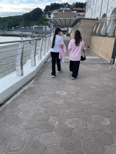 Two young women are walking along a pedestrian walkway in Olginka, Russia. One woman is wearing a white shirt and black pants, while the other is wearing a pink shirt and black pants. They are walking parallel to a railing that overlooks the coast, including the sea and forested hillside. The walkway is paved with patterned gray stones. In the background, buildings and trees can be seen on the hillside.