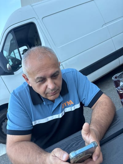 An over-the-shoulder shot shows a middle-aged man with grey hair looking down at his phone. He is wearing a blue polo shirt with black accents and orange lettering that reads "DYNA." The man is seated at a dark picnic table, and in the background, a white cargo van is parked. The scene appears to be outdoors, possibly in an industrial or commercial area, consistent with locations in the city of Boom, Belgium, known for its industrial heritage and large event venues.