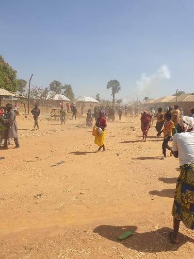 A wide, dusty ground stretches under a bright sky, holding a scattered crowd. People, some in vibrant traditional attire, move amidst structures suggesting a village setting. A tall tree stands out, with a faint smoke trail nearby. The ground is arid and light brown, dotted with sparse vegetation and debris. Shadows stretch from the figures, indicating a sunlit environment.