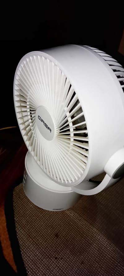 The image displays a white electric fan, viewed from a slightly elevated side angle. The fan features a modern design with a circular grille composed of numerous thin, radial white plastic fins. The brand name "QASA" is clearly visible in black letters on the central hub of the fan's front grille. The fan unit is predominantly white plastic, including its base and the curved support arm connecting the fan head to the base.

The fan is placed on a textured surface, which appears to be a brown-and-tan woven rug or mat, parts of which are visible in the foreground and beneath the fan's base. The background is dark, suggesting low light conditions or a plain dark wall/backdrop behind the fan. There are no people visible in the image, and no specific activity is taking place; the fan appears to be stationary. The scene depicts an indoor setting, likely a room in Rijiyar Zaki, Nigeria.