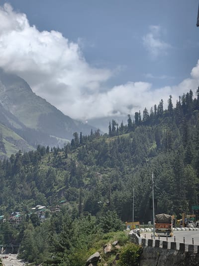 The image shows a panoramic view of a mountainous landscape near Kulang, India. The foreground features a winding road with a truck and several vehicles, bordered by a low stone wall and vegetation. Further back, a cluster of buildings, likely a village or town, is nestled amidst lush green trees. The mid-ground is dominated by a densely forested mountainside, with a variety of evergreen trees reaching towards the sky. The background reveals towering, majestic mountains partially obscured by clouds, creating a dramatic and atmospheric scene. The sky above is a mix of blue and white clouds, with the sun casting shadows and illuminating parts of the landscape. The overall impression is one of natural beauty and a remote, mountainous setting.
