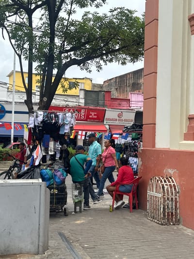 This image depicts a street scene in Perímetro Urbano Barranquilla, Colombia. In the foreground, a grey rectangular object, possibly a public utility box or planter, is visible on the left. Further back, a black shopping cart is laden with assorted items, including large dark plastic bags and two colorful bundles. A man in a green shirt and dark pants is seated on a stool to the right of the cart, with his back to the camera.
In the mid-ground, a clothing rack is displayed with various items of clothing, predominantly socks, hanging on it. Several flags, including the flags of Norway, Australia, and the United Kingdom, are visible on poles to the left and right of the clothing display. People are browsing or interacting around the stall. A man in a red shirt is standing to the left, and a man in a blue plaid shirt and a man in a light blue shirt are conversing to the right. A woman in a pink shirt and jeans walks past, looking upwards. Another woman in a red top and denim shorts sits on a red plastic chair on the far right, facing away from the camera.
In the background, buildings of various colors and heights are visible, including a bright yellow building and a pink building. A large tree with dense green foliage partially obscures the sky. The ground is paved with brick-like tiles. To the right of the woman in the red top, a decorative white metal cage-like structure is attached to a coral-colored wall. The sky appears overcast.