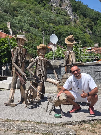 In the village of Golyamo Belovo, Bulgaria, a man wearing a white t-shirt and white shorts, with sunglasses and a watch, crouches next to a wooden sculpture of a dog. He is giving a thumbs up. Behind him are three wooden figures resembling people, adorned with straw hats, and a wooden spinning wheel. In the background, houses with red tile roofs and lush green trees cover a rocky hillside. The scene is set on a paved outdoor area with scattered rocks.