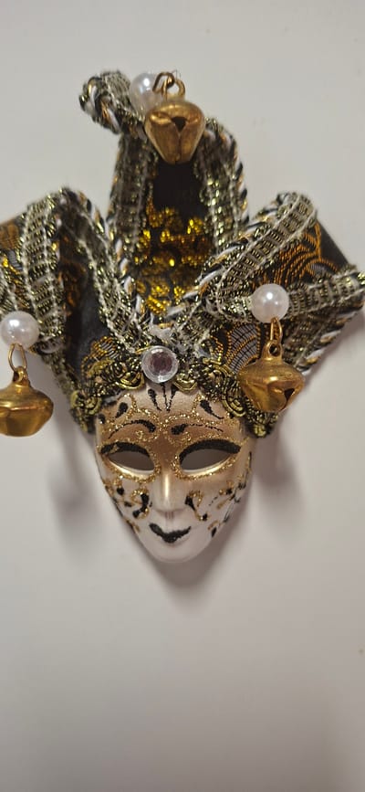 The image shows a close-up, straight-on view of a decorative mask. The mask is designed in a jester or harlequin style, with a three-pointed hat adorned with gold bells and pearls. The mask itself is predominantly gold with black accents and glitter detailing around the eyes and across the forehead. A small, clear gem is placed above the nose. Black lines create a stylized facial expression, with dark, empty eye sockets and a small, downturned mouth. The mask is positioned against a plain white background. There is no indication of the city of Orimattila, Finland, in the image or its context.