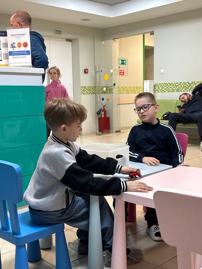 The photograph captures an indoor scene, possibly in a communal space like a cafeteria or waiting room, though the exact location in Kuleshovka, Russia, is uncertain without additional context. Several individuals are present, some seated at tables and others in the background. A boy wearing a gray and black jacket sits at a blue chair, playing with a small red toy car on the table. Across from him, another boy in a dark track suit with white stripes sits with his hands on the table, looking towards the boy with the car.

In the background, other people are present. One individual is seen lying down on a green bench with their head propped up. Near the front of the scene, another person is seen sitting near a sign and a woman with glasses can be seen peering through a turquoise paneled wall.

The environment includes elements like colorful chairs and tables, suggesting a child-friendly area. A fire extinguisher is visible on the wall. A sign, partly obscured by the boy sitting, contains some printed text in Russian. The location appears to be well-lit.