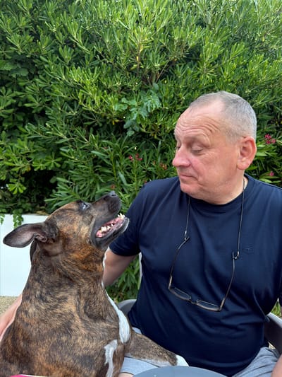 In Općina Baška, Croatia, a man with short grey hair and a navy blue t-shirt is looking down at a brindle-colored dog. The dog is looking up at the man with its mouth slightly open, revealing its teeth and tongue. The man has a pair of glasses hanging around his neck. They are sitting outdoors with lush green foliage behind them. The man's right hand is resting on the dog's back.
