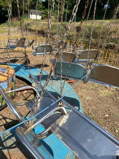 This is a photograph of an empty swing ride at an amusement park, possibly located in Karangasem, Indonesia. The ride consists of multiple chain-hung seats, with some showing signs of rust. The ground is dirt and dry grass, with some loose rocks. In the background, there is a yellow barrier fence and a small white building situated on a grassy hill. The sky is clear and blue, suggesting a sunny day. The focus is on the ride's structure, with the seats appearing empty and unused.