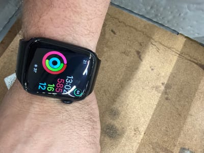 A close-up, elevated, eye-level, medium shot shows a person's left wrist wearing a black Apple Watch Series 7. The watch face displays the time "13:02", activity rings (red, green, and blue), and data points "31°", "21", "12", "16", "585", and "64". The watch is positioned on a textured, light brown, wooden surface, possibly a workbench or desk. The background consists of a grey, textured surface and a soft shadow cast across the wooden surface. The image is in focus on the watch and wrist. No specific location in Chociule, Poland is identifiable.