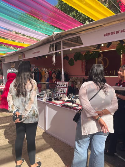 The image shows a market stall in Chandigarh, India, located at the coordinates 30.764 latitude and 76.798 longitude.  A woman in a white floral blouse and black pants is looking at the display of necklaces and jewelry on a table inside the stall. She is holding a cell phone in her left hand.  Another woman, wearing a white embroidered top and blue jeans, is standing behind her, looking at the jewelry.  There is a third person in the background, a young woman wearing a red top and holding a tray of jewelry.  The stall is decorated with pink and white fabric and has a sign that reads "ABBREVA BY ROS".  The stall is situated under a canopy of colorful fabric that is strung overhead. The ground is dirt.  The sun is shining brightly.