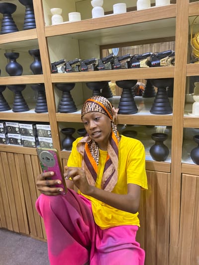 In Unguwar Sarki, Nigeria, a young woman wearing a yellow t-shirt and a pink and purple skirt sits in front of a wooden shelf displaying various items. She is holding a smartphone in her left hand and appears to be interacting with it. The shelves behind her are filled with an assortment of objects, including what appear to be small, dark vases or decorative pieces, boxes labeled "DONUT CHARCOAL," and several black, intricate candle holders. Further up the shelves, there are white ceramic containers and a row of what seem to be tools or small machinery, possibly for charcoal production. The lighting suggests an indoor setting, possibly a shop or storage area.