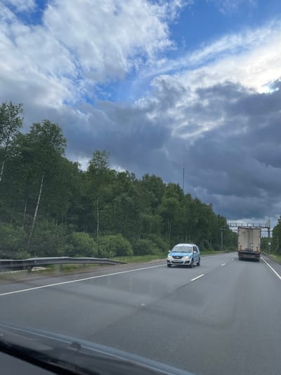 The image shows a highway near Novyi Byt, Russia. A light blue and white car and a truck are traveling on the wet asphalt road, alongside a dense forest under a cloudy sky.