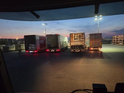 The image depicts a cargo loading area at Frankfurt Airport (FRA) in Germany, during dusk. A fleet of four semi-trailers is parked on the tarmac. The sky displays hues of orange, pink, and blue, indicative of the twilight hours. Tall industrial lamps illuminate the scene. In the foreground, the interior of a vehicle's cabin is visible, suggesting the photo was taken from a driver's perspective.
The first trailer from the left is a white box truck with a red stripe and a logo that appears to be "K+H Trailer." To its right, another white box truck is visible, with what appears to be cargo wrapped in plastic sheeting stacked on pallets next to it. The third trailer, also a box truck, is partially open, revealing its internal structure. The fourth trailer, a flatbed, is also partially open, showing its loading area.
A person wearing a yellow safety vest and dark pants is standing near the first trailer, seemingly overseeing operations. The ground is paved with asphalt and shows some reflective wetness, possibly from recent rain or cleaning. In the background, the airport's infrastructure, including buildings and what appears to be a distant airplane, can be observed. The overall atmosphere suggests an operational logistics environment at the airport.