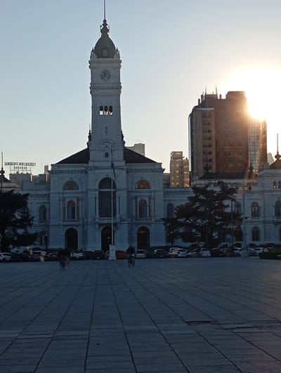 The image depicts the front of the Government Palace (Palacio de Gobierno) in La Plata, Argentina, bathed in the warm light of a setting sun. The grand, white neoclassical building features a prominent clock tower with a dark dome and a spire. In front of the palace, a large paved square is visible, with several cars parked along the street in the background. Two individuals are seen on bicycles in the square. The sky is clear and light blue, with the sun creating a bright halo in the upper right quadrant of the image. The overall scene conveys a sense of civic grandeur and tranquil urban life.