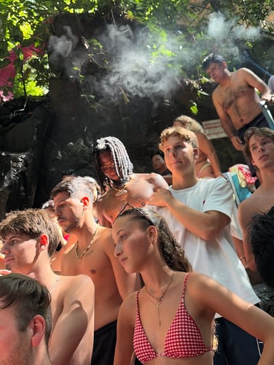 In Gianyar, Indonesia, a group of individuals stands in a humid, natural setting, likely near a waterfall or hot spring, evidenced by the visible steam rising in the background. The scene is crowded, with many people visible, most of whom are young adults. The lighting suggests an outdoor, daytime environment, with sunlight filtering through dense foliage and casting shadows.

In the foreground, on the left, a young man with short, light brown hair is partially visible. To his right, another young man with darker hair and a beard is looking to the side, wearing a necklace. Behind them, a person with dreadlocks and glasses is also looking down.

In the center of the image, a young man with blond hair and a white t-shirt is looking forward, with his hand resting on another person's shoulder. To his right, a young man with light brown hair is looking away.

Further back, a man with dark hair and a tan torso is visible, looking towards the right. In the foreground right, a young woman with long, curly brown hair is looking to the side, wearing a red and white checkered bikini top. A second young woman with dark hair is seen in the far right.

The environment is lush and green, with trees and rocks forming the backdrop. The steam suggests a geothermal activity or a misty atmosphere from a water source. The overall impression is of a group enjoying a natural tourist attraction.
