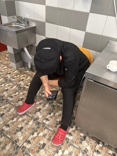 An individual is depicted seated indoors in what appears to be a utility or backroom area within Kirkkolakhti, Russia. The person, a male, is wearing a black long-sleeved work jacket, black pants, a black baseball cap worn backward, and red Crocs with white soles. He is positioned with his head bowed, leaning forward, and holding a dark-colored smartphone horizontally in his left hand, observing its illuminated screen. He is seated on a light brown wooden stool, partially visible behind him. The floor is covered with patterned tiles in shades of brown, beige, and dark grey. The walls are tiled with a checkerboard pattern of alternating grey and white square tiles. To the left, a wall-mounted stainless steel utility sink with a faucet is installed, featuring the "КАМИК" (KAMIK) brand name. A white flexible drain hose extends from beneath the sink. A segment of red flooring or a mat is visible in the lower left corner. To the right, a stainless steel countertop and cabinet unit are present, with a white ceramic mug resting on the countertop.