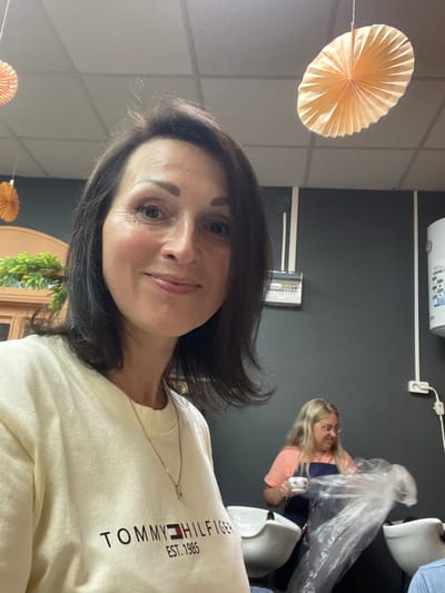 The media file shows a selfie taken by a woman with dark hair. She is wearing a light-colored t-shirt with the text "TOMMY HILFIGER EST. 1985" visible on it. She is also wearing a delicate gold chain. Her face is turned towards the camera, and she has a slight smile. Her eyes are brown. In the background, another woman with blonde hair is visible, wearing an apron and holding a roll of plastic wrap. She appears to be in a salon or a similar service setting. There are white chairs and a dark wall behind her. To the left of the main subject, a decorative paper fan in a peachy-orange color is hanging.
The scene is indoors, as indicated by the artificial lighting and the presence of furniture and equipment. The time of day is not explicitly indicated. The presence of decorative elements like the paper fan suggests a festive or welcoming atmosphere. The overall mood seems relaxed and informal. The location context provided is Кумёны, Russia, but there are no explicit visual cues within the image to confirm this specific location.
