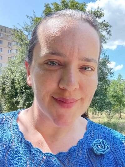 A woman, likely in her late 30s to early 40s, is taking a selfie outdoors. She has fair skin with some visible pores and fine lines. Her hair is dark brown and pulled back. She is wearing a blue, crocheted top with an open knit pattern. A small, blue crocheted flower with a light-colored gem in the center is attached to the left side of her top. Her left ear has a red stud earring.
The background suggests an urban environment with residential buildings visible behind trees. The sky is bright blue with scattered white clouds. The lighting indicates it is daytime and sunny. The setting appears to be a park or residential area with green trees and some dry grass or brush. It is possible this photo was taken in Kherson, Ukraine.