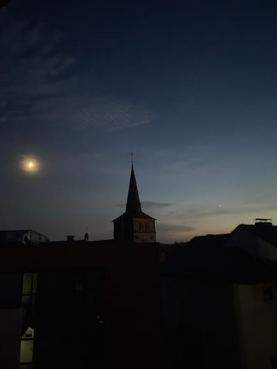 The image depicts a nighttime scene in Waldbröl, Germany, featuring a prominent church steeple silhouetted against a dark sky. A crescent moon is visible in the upper left quadrant, casting a soft glow. The foreground is dominated by dark rooftops and the facade of a building, with some windows showing faint interior light. The sky transitions from a deep blue at the top to lighter hues near the horizon, suggesting twilight or early dawn. The overall atmosphere is quiet and serene.