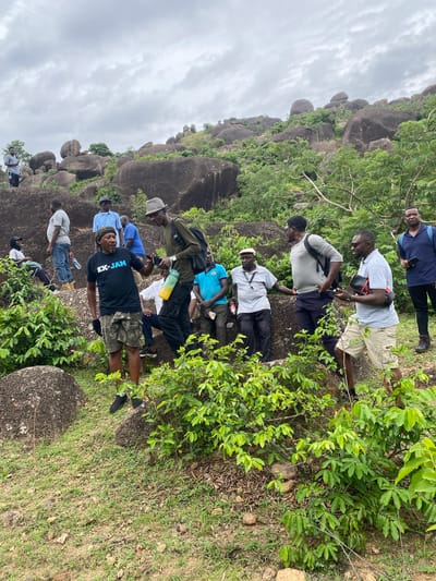 The image shows a group of about 10 people standing on a rocky, grassy hillside. The location is likely in or near Ganawuri, Nigeria, given the query context. The hillside is covered with large boulders and green vegetation. The sky is overcast. The people are of African descent and are dressed in casual clothing. Some are looking at their phones or cameras. The overall impression is that the group is hiking or exploring the area.