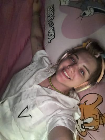 A young woman with fair skin, blue eyes, and a bright smile lies in bed. She is wearing gold headphones and a white, short-sleeved pajama top with a black "V" on the pocket. She also wears a green beaded necklace. She is positioned between two pink Tom and Jerry-themed pillows. The scene seems to be indoors, possibly in Большая Шильна, Russia, but there are no location-specific details in the image to confirm. The subject is taking a selfie.