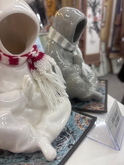 Two ceramic figures, one white and one gray, are seated side-by-side on decorative mats. The figures have hooded heads and appear to be wearing scarves. The white figure has a red and white striped scarf with white fringe, while the gray figure has a gray and white checkered scarf. The white figure's hands are clasped in front of its body. Both figures are positioned on what appears to be a white surface, possibly a table or shelf. In the foreground, a clear plastic price tag holder is visible, displaying Korean text and the price "₩42,000". In the background, out-of-focus items are visible, including what appear to be more decorative objects and possibly some artwork. The overall impression is that of items displayed in a shop or market setting. The presence of the price tag in Korean indicates this is likely in South Korea, and given the context of souvenir shops and cultural items, a location in Gyeongju-si, a city known for its historical and cultural significance in South Korea, is plausible.