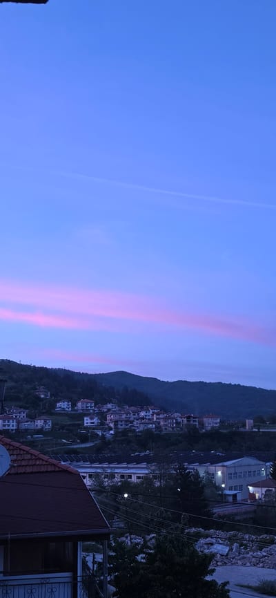 The image shows a view in Rudozem, Bulgaria, at dusk or dawn. The sky is a gradient of blue to pink, with distinct pink bands. A mountain range separates the sky from a cluster of houses and buildings along the slope. A portion of a roof is visible in the lower right corner, suggesting the photograph was taken from an elevated position. Streetlights are visible within the residential area. Overhead power lines are present.