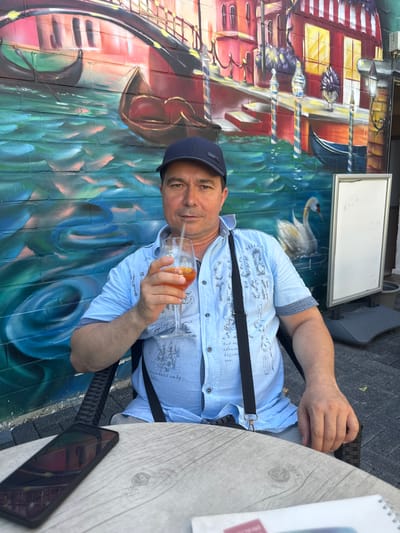 A man wearing a blue baseball cap and a light blue patterned short-sleeved shirt sits in a chair facing the camera, holding a glass of orange liquid with a straw in his right hand. He is seated at a wooden table with a smartphone on it. Behind him is a vibrant mural depicting a Venetian canal scene with gondolas and buildings. To the right of the man, a white signboard on a stand is visible. The man appears to be at an outdoor seating area of a restaurant or cafe in Wolfsburg, Germany.