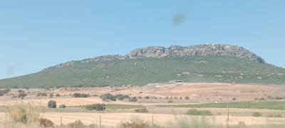The image shows a wide, hilly landscape under a clear blue sky. In the background, a large mountain range with a rocky, jagged crest is visible. The slopes of the mountain are covered in dense, green vegetation, giving way to sparser vegetation and bare earth on the lower hills. In the middle ground, there is evidence of construction or earthmoving activity, with large piles of dirt and some rudimentary structures. A yellow bulldozer is visible on the disturbed earth. The foreground consists of fields of dry, yellow grass, interspersed with scattered trees and some green patches of vegetation. The overall scene suggests a rural or semi-rural area, possibly on the outskirts of a city, with a mix of natural and developed landscapes. There are no people visible in the image. The presence of construction equipment indicates ongoing development or alteration of the land.