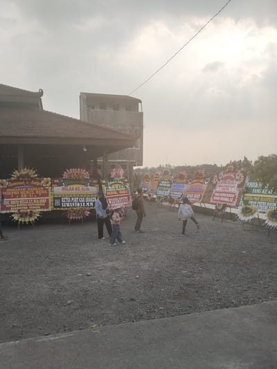 The image captures an outdoor scene in Plosorejo, Indonesia, likely during daylight hours with overcast skies. The primary focus is a collection of large floral arrangements, commonly known as "papan bunga," displayed in front of a building with a traditional Indonesian roof. These arrangements are adorned with congratulatory messages and names, indicating a celebration or commemoration.

Several people are present, mingling and observing the floral displays. In the foreground, a woman and a young child are visible, with the child appearing to interact with the display. Further back, other individuals are seen standing and walking around the area. The ground is covered in gravel and dirt, typical of an outdoor gathering space.

The visible text on the floral arrangements suggests an event celebrating the 41st anniversary of "PONPES NURUL HUDA" (Nurul Huda Islamic Boarding School). Some of the messages also mention "MILAD KE-41 th" (41st Birthday) and include names such as "ERIC HOSTA MELLA, ST., M.SC. QRMP KEPALA KANTOR PERTANAHAN KABUPATEN SRAGEN" (Head of the Land Office, Sragen Regency), "SUWANTO.S.E.M.M KETUA PSHT CAB SRAGEN" (Chairman of PSHT Sragen Branch), "WAKIL BUPATI SLEMAN DANANG MAHARSA, SE" (Deputy Regent of Sleman, Danang Maharsa, SE), and "NOVITA PUT HASTUTI INDAH PUTRI BATIK". These indicate the presence of local officials and organizations. The overall atmosphere appears to be one of a formal but celebratory gathering. The background shows a building structure and some greenery, suggesting a semi-urban or rural setting.