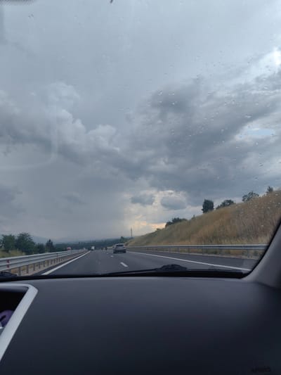 This media file shows a view from inside a car driving on a highway. The scene is a highway with multiple lanes, a metal guardrail, and a grassy embankment on the right side. In the distance, there are trees, hills, and some buildings. The sky is overcast with dark, heavy clouds, suggesting a stormy or rainy day. There are raindrops visible on the car's windshield.
On the highway, several vehicles are visible, including cars and what appear to be motorcycles. One car is in the right lane, and other vehicles are further ahead. The overall mood is somewhat somber due to the weather conditions. The time of day appears to be daytime, given the visibility, but the cloudy sky creates a dim light.
The location context provided is Dolni Bogrov, Bulgaria, which fits the description of a highway scene with natural surroundings. There is no visible text in the image. The activity is driving on a highway. The subjects are vehicles, the highway, and the surrounding landscape under a cloudy sky.