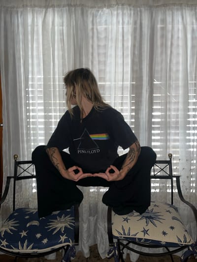 A person sits on a chair in front of a window. The person is wearing a black t-shirt with "Pink Floyd" printed on it and black pants. The person is making an "OK" sign with both hands. The chair has a blue and white patterned cushion. The window has white curtains and blinds. The location is inside a building. There is a light floral patterned cloth on the floor. The location is unspecified and therefore cannot be linked to the city of Saladillo, Argentina.