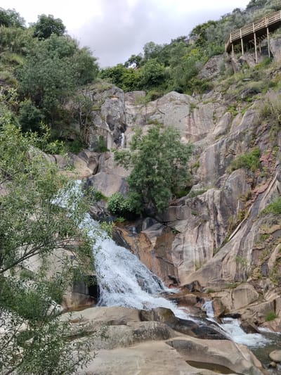 The image depicts the Fisgas de Ermelo waterfalls in Alijó, Portugal. Water cascades down a rocky terrain, with vegetation clinging to the sides. A wooden walkway is visible at the top right, suggesting a maintained path for viewing the falls. The sky is overcast. The foreground features trees, partially obscuring the view of the waterfall.