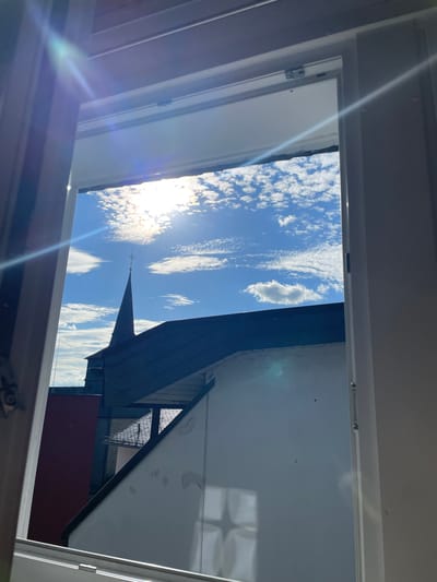 The image shows a view looking out of an open window or skylight, with a bright blue sky dotted with fluffy white clouds. The sun is shining brightly, creating lens flares across the image. In the middle ground, a dark, steep roof of a building is visible, with a thin spire or steeple extending upwards towards the sky. Part of a red wall is also visible. The foreground shows the interior frame of the window or skylight, with some wooden elements and metal hardware. The overall scene suggests it is daytime, with clear and sunny weather. The presence of the spire suggests a building with some architectural significance, possibly a church or historical structure, located in Waldbröl, Germany. The view is angled and appears to be taken from an elevated position.