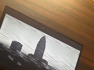 A damaged laptop screen displays a black and white image of a cityscape. The architecture appears to depict the Key Tower in Cleveland, Ohio, not Eneka, Nigeria. The image on the screen is distorted by horizontal lines, indicating damage to the display. The laptop rests on a wooden surface.