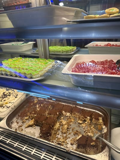 The image shows a display case filled with various desserts, likely in a bakery or ice cream shop. The primary subjects are the desserts themselves, including what appears to be a tray of tiramisu with a serving utensil, a container of small, square green pastries, and a tray of red jelly-like candies. There are also some pastries or cakes visible on the left side of the frame, some with yellow frosting and a green filling. The setting is indoors, with glass panes reflecting the interior lights and some of the surrounding environment. The lighting is artificial, casting a slightly cool tone. The display case appears to be refrigerated, with visible metal grates on the right side of the frame, suggesting it's a food service establishment. There is no visible text in the image. The image is taken from a slightly elevated angle, looking down into the display case. The activity suggested is the display and potential serving of desserts. The desserts appear to be freshly made or recently served, with some being partially eaten. The overall impression is one of a well-stocked dessert display.