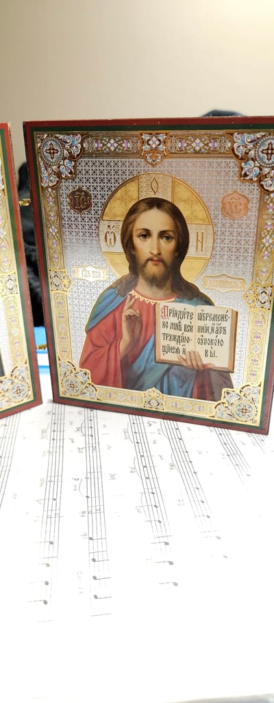 The image shows an icon of Jesus Christ, likely an Eastern Orthodox icon, placed on a white surface with handwritten musical notation. Jesus is depicted with a halo, holding an open book in his left hand and making a blessing gesture with his right hand. The icon has intricate decorative borders with floral and geometric patterns, rendered in gold, enamel, and other colors. The text on the book and beside Jesus is in Cyrillic script. The musical notation appears to be handwritten, with staff lines and notes, and some additional markings that could be lyrics or performance instructions. The overall scene suggests a quiet, possibly devotional or artistic setting, with the icon and music sheets placed together. The lighting suggests it is indoors. There are no specific cues for time of day or weather.