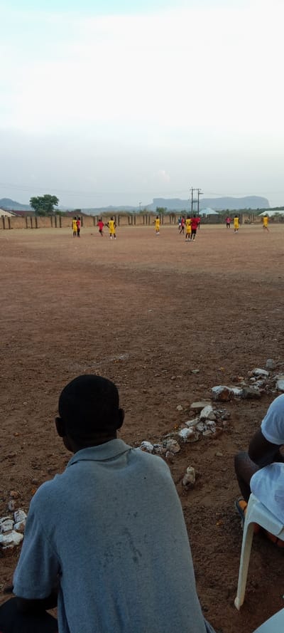 People in red and yellow uniforms play soccer on a dirt field bordered by a wall and greenery. Overhead power lines stretch across the sky. In the foreground, the back of a person in a gray shirt is visible, sitting on the ground next to a row of painted white rocks. Part of another person is visible to the right of the image sitting in a white plastic chair.