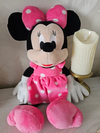 A plush Minnie Mouse toy lies on a textured surface, possibly a bedspread or couch. The toy is primarily pink and black, with white polka dots on its ears and dress. Its eyes are closed, giving it a serene or sleepy appearance. To the left of the toy, an artificial candle with a gold base is placed on its side. The candle is a creamy white color and appears to be made of wax. The background is a light, neutral color with a subtle pattern. The scene is brightly lit, suggesting daytime indoors. There is no visible text in the image.