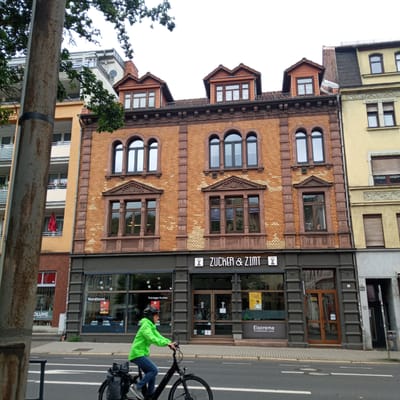 The image shows a street scene in Eisenach, Germany, featuring a brick building with several businesses on the ground floor. The building has a traditional architectural style with arched windows on the upper floors. On the ground floor, there is a shop labeled "ZUCKER & ZIMT" with large glass windows and an entrance. To the left of this shop, another establishment is partially visible with signage that includes "OLUME HAARSTUDIO" and "Konditorei Thüringer Kuchen." To the right of "ZUCKER & ZIMT," a sign advertises "Eiscreme Hausgebackene Waffel." A person wearing a bright green rain jacket and a helmet is riding a bicycle in the foreground, on the street. The street is paved and has road markings. The environment appears to be a commercial area in a German city.